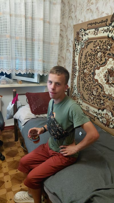 A young man with short, light brown hair, wearing a green t-shirt, maroon shorts, a black watch, and a black fanny pack across his chest, sits on a bed in what appears to be a room in Borodianka, Ukraine. He is holding a clear glass with a dark liquid in his right hand and has his left hand on his hip. The room has a window with a white lace curtain to the left, a patterned rug on the wall to the right, and a dark red pillow on the bed behind him. The floor is tiled with a brown and cream checkerboard pattern.