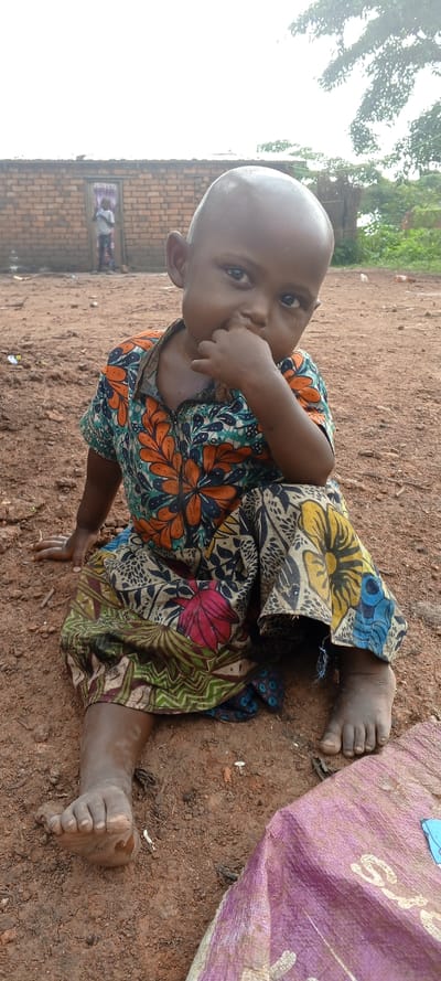 The image captures an outdoor scene in Kakungu, Democratic Republic of the Congo. A young child with dark skin and a shaved head is seated on reddish-brown earth in the foreground. The child wears a short-sleeved garment with a vibrant pattern of orange, blue, green, yellow, and red floral and geometric motifs. The child's right hand is raised, with fingers touching the mouth, and both bare feet are visible, coated with dirt. The child looks directly forward. In the background, a building constructed of exposed reddish-brown bricks features a doorway draped with a patterned fabric. A second child, wearing a light-colored top and patterned lower garment, is partially visible standing within this doorway. Sparse vegetation and trees with green foliage are present in the far background, beneath an overcast sky. A crumpled purple woven sack lies partially visible in the bottom right corner of the frame.