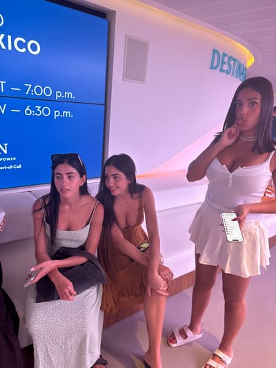 Three young adult females are depicted in an indoor waiting area, likely a port lounge or ferry terminal, in San Miguel de Cozumel, Mexico.
To the left, a female with long dark hair, wearing a light green polka-dot maxi dress and sunglasses pushed onto her head, sits on a white padded bench. She holds a black bag in her lap and a white smartphone in her left hand. Her facial expression is neutral, and she looks slightly to her left.
Adjacent to her, another female with long dark hair, wearing a brown sundress, sits on the same white padded bench. She is smiling and looking towards the female on her left. A yellow smartphone rests on the bench next to her right thigh.
To the right, standing, a third female with long dark hair, wearing a white sleeveless top with lace trim and a white pleated mini-skirt, faces the camera. She holds a white smartphone with its screen visible in her left hand and gestures towards her chin/neck area with her right hand. Her expression is playful, with her tongue slightly extended and lips pursed. She wears a pearl necklace and mismatched sandals (white double-strap on the left foot, light-colored double-strap on the right).
In the background, a large blue digital display screen shows text including "MEXICO", "T – 7:00 p.m.", "V – 6:30 p.m.", "POWER", and "Port-of-Call", suggesting departure information. Above the screen is a white speaker grille. The walls are white and feature a gentle wave-like design, illuminated with a soft magenta hue. A partially visible blue sign on the right wall reads "DESTIN". The flooring is light gray.