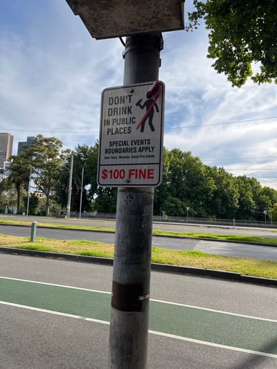 The image displays an urban outdoor scene on a clear, sunny day, with a metal pole featuring a prominent sign in the foreground. The sign is white with black text and a red graphic, depicting a stylized figure holding a bottle with a red strike-through, prohibiting alcohol consumption.
The text on the sign reads:
"DON'T DRINK IN PUBLIC PLACES"
"SPECIAL EVENTS BOUNDARIES APPLY"
"New Years, Moomba, Grand Prix Events"
Below this, in red, it states: "$100 FINE". A small number "76" is also visible on the pole.
In the background, lush green trees line what appears to be a park or a treed median strip. Beyond the foliage, a multi-lane road is visible, featuring a distinct green-painted lane, typically used for bicycles. The sky is bright with scattered white clouds. In the far distance, faint outlines of urban buildings suggest a city skyline. The presence of the sign, particularly referencing "New Years, Moomba, Grand Prix Events," and the provided context, indicate the location is likely a public area in Southbank, Australia, a city known for hosting such large-scale events. The scene is devoid of people or direct activity, but the sign itself serves as a regulatory notice regarding public behavior.