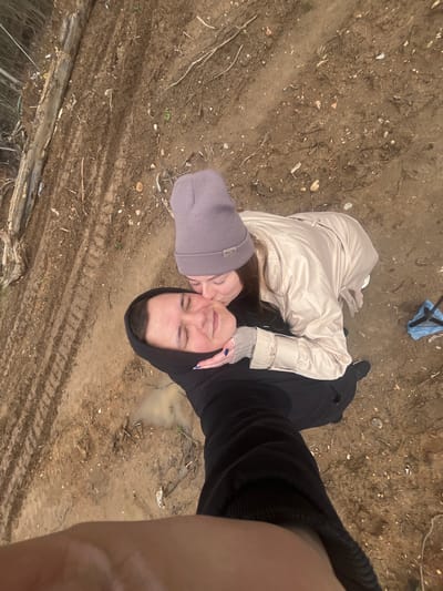 Two individuals are posing for a selfie in what appears to be an outdoor environment in Ivlevo, Russia. The ground is predominantly dirt, showing signs of tire tracks and some puddles. One individual is wearing a lilac beanie and a light-colored coat, appearing to kiss the cheek of the other individual, who is wearing a black hooded sweatshirt. A blue bag with an unidentified object is present in the background.