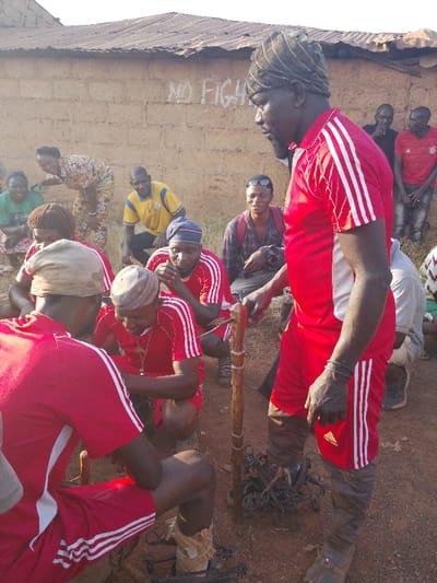 The scene depicts an outdoor gathering on reddish-brown dirt ground in Vodni, Nigeria, featuring approximately ten to fifteen individuals. In the background, a light brown mud-brick building with a corrugated metal roof is visible. The phrase "NO FIGHT" is spray-painted in white on the building's wall.

In the foreground, a tall, dark-skinned man stands, facing left and slightly downwards. He wears a red athletic shirt with white Adidas stripes, matching red shorts, and dark brown leg coverings or boots. A dark fabric is wrapped around his head. He holds a slender, carved wooden stick vertically with his right hand; the stick has several small, sharp metal tines protruding from its upper section.

Around the base of this stick and near the feet of the standing man, a pile of dark metal objects, resembling animal traps or snares, rests on the ground. Three other men, also wearing identical red athletic shirts with white stripes, are crouched or kneeling in front of the standing man, intently focused on the objects. One crouches with his back partially to the viewer, wearing a light-colored head covering and dark leg coverings similar to the standing man's. Another, wearing a blue and white patterned head covering, rests his hands near his face. The third, wearing a light-colored cap and a metallic pendant, appears to be actively manipulating an object on the ground.

In the mid-ground and background, several other individuals observe the central activity. These include a woman in a patterned yellow dress bending over and smiling, a man in a yellow polo shirt, a man in a plaid shirt with sunglasses on his head and a red backpack, and several other men, some standing, some partially obscured, wearing various casual attire including red and black shirts. The overall impression is one of a community engaged in an instructional or practical demonstration involving the wooden stick and metal traps.
