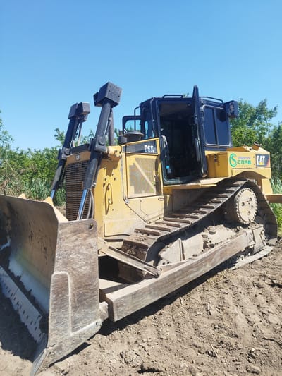 A yellow Caterpillar D6R2 bulldozer is parked on a dirt construction site. The bulldozer is equipped with a large front blade and has a black cab with tinted windows. The doors of the cab are open, revealing the interior. The machine is positioned at an angle, with the blade closest to the viewer. In the background, there are trees and green foliage under a clear blue sky, suggesting a sunny day. The ground is covered in dirt and some dry grass, indicating an outdoor, possibly rural, setting. There is visible text on the bulldozer, including the "CAT" logo, "D6R2", and a circular green logo with the text "СЛАВДОРОЙ" and "СПАВ" in Cyrillic. The scene appears to be in Rozdil, Ukraine. No people are visible. The overall impression is of heavy machinery at a construction or land-clearing site.