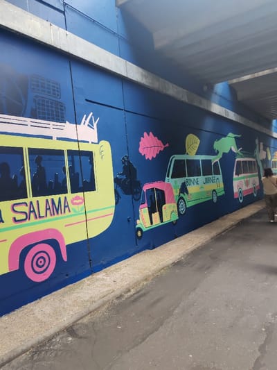 This image depicts a section of a concrete underpass wall in Fontenay-sous-Bois, France. The wall is painted deep blue and features a multi-colored mural of stylized vehicles and figures. From left to right, the mural includes: a large lime-green bus with pink accents and a white roof rack, displaying "SALAMA" in white text and containing dark blue silhouetted passengers; a dark blue silhouette of an individual on a bicycle; a pink and lime-green three-wheeled auto-rickshaw; a lighter green bus with "BONNE JOURNEE" in white text and silhouetted passengers; and a partial view of a pink and white striped vehicle with a white roof rack. Above the vehicles, painted elements include a pink leaf, a yellow-green leaf, and a green feline-like creature. The ground consists of a light grey concrete curb separating the wall from a dark grey asphalt path exhibiting minor discoloration. A single individual, observed from behind, walks towards the right along the asphalt, wearing a light-colored top and dark, patterned trousers. The underpass ceiling, visible at the top, is constructed from grey concrete.