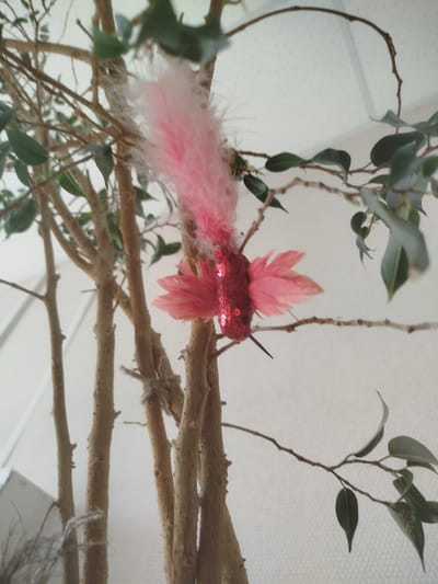 A decorative hummingbird ornament, made of red sequins and pink feathers, is attached to a branch of a potted tree. The tree has multiple brown trunks and green leaves. The background is a textured white wall. The location is not specified in the image or description.