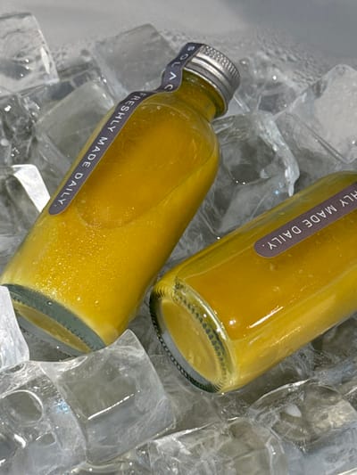 Two glass bottles filled with yellow liquid, possibly juice, rest on a bed of ice. A label reading "FRESHLY MADE DAILY" wraps around each bottle. One bottle displays a silver cap with "SOLACE" visible on a sticker. The clear ice cubes reflect light, creating highlights on the bottles and surrounding surfaces. The close-up shot emphasizes the chilled and refreshing nature of the beverage.