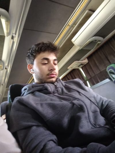 The image depicts the interior of a passenger transport vehicle, likely a bus or coach. The precise location within the city of Mankat al-Hatab, Syria, cannot be determined from the visual evidence provided.
A young male individual, appearing to be in his late teens or early twenties, is seated in a dark-colored passenger seat. He has short, dark, disheveled hair, a light mustache, and a short beard. He is wearing a dark grey, full-zip hooded sweatshirt with drawstrings at the hood. A white wireless earbud is visible in his left ear. His head is tilted slightly forward, and his gaze is directed downwards, out of frame.
The vehicle's interior features a dark grey ceiling panel. Along the upper periphery, multiple white rectangular light fixtures are visible, some with a yellow strip running parallel to them. Dark brown pleated curtains cover windows along the right side of the frame. A small, white rectangular fixture, possibly an air vent or reading light, is mounted above the curtains. A partial view of another seat with light-colored fabric is visible to the subject's left, out of focus. No other individuals or explicit interactions are visible within the frame.