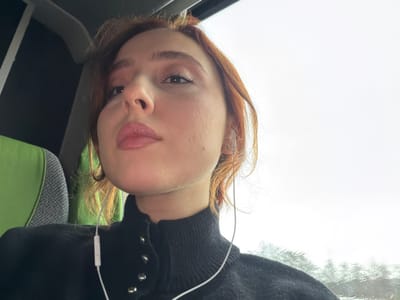 The image depicts a young light-skinned female in a low-angle close-up within a vehicle, located in Moineville, France. She has reddish-brown hair, styled loosely, and light makeup on her eyes and lips. She wears a black high-neck top with several silver-toned snap buttons visible on the collar area. White wired earphones are inserted into both ears, with the wire draped across her left shoulder and chest. Her gaze is directed slightly upwards and to the viewer's right. To the left, behind her, a portion of a vehicle seat with green and grey upholstery is visible. To the right, through a window, an overcast sky is visible, with streaks of water indicating rain on the glass surface. The interior of the vehicle has dark overhead elements. No other individuals are present.