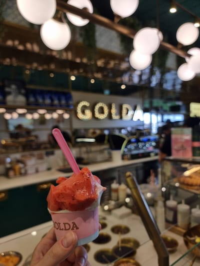 A close-up, canted image captures a cup of strawberry gelato held by a hand, with a pink plastic spoon inserted. The cup has the word "GOLDA" printed on it. The backdrop is a blurred interior of a gelateria, with visible display cases containing various flavors. Further back, the word "GOLDA" is spelled out in large, illuminated letters, matching the brand name. Soft, globular lights hang on the left. Based on the brand, this scene is likely taking place at a Golda ice cream shop in Bnei Shimon Regional Council, Israel.