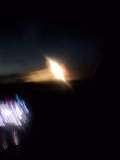 The image depicts a blurry, abstract scene at night, possibly taken from a moving vehicle. In the upper center, a bright, rectangular light source is visible, suggesting a building or a very strong artificial light. The light is diffused and has a warm, yellowish-orange hue, with streaks of orange light emanating from its right side. Below this light, the landscape is dark and indistinct, with some textured areas that could represent clouds or terrain. In the lower left quadrant, there are streaks of colorful lights, predominantly white and blue, with some hints of red and orange. These lights are elongated and blurred, consistent with motion blur from vehicle headlights or taillights. The overall impression is one of movement and a low-light environment. There is no specific information about the city of Yettitom, Uzbekistan, that can be inferred from this image.