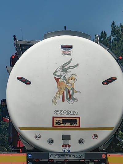The media file shows the rear of a white tanker truck. The truck has a large, round white tank with various decals and markings. Dominating the center of the tank is a cartoon image of two characters, resembling Bugs Bunny and Lola Bunny from the Looney Tunes franchise, engaged in a sexually suggestive act. Bugs Bunny is depicted spanking Lola Bunny.

Above the cartoon, there are several logos and stickers. One prominent sticker contains the word "SCANIA" in large letters. Below "SCANIA," there is a red rectangular sticker with a depiction of a truck and trailer.

On the left side of the truck's tank, there is a set of three red lights arranged horizontally. On the right side, there is a similar set of lights, with the top one being red and the bottom two appearing to be amber or orange. There are also smaller red lights visible on the side of the truck's cabin on the left.

Towards the bottom of the tank, there are circular stickers. One on the left reads "BG," likely indicating Bulgaria as the country of origin or registration. To its right, a black circle contains the number "60." Further to the right, another black circle contains the number "80." A circular red and white sticker on the right displays the number "90" within a red circle, indicating a speed limit.

At the very bottom of the image, a metal bar with red lights is visible, and a company name, "CISTERNY.COM," is printed on it. The sky above is clear and blue, suggesting it is daytime with fair weather. The background shows green trees, indicating an outdoor setting, likely on a road or highway.

The overall scene captures a large vehicle with a provocative and humorous sticker, along with official markings and company information. The location context is given as Malinovo, Bulgaria.