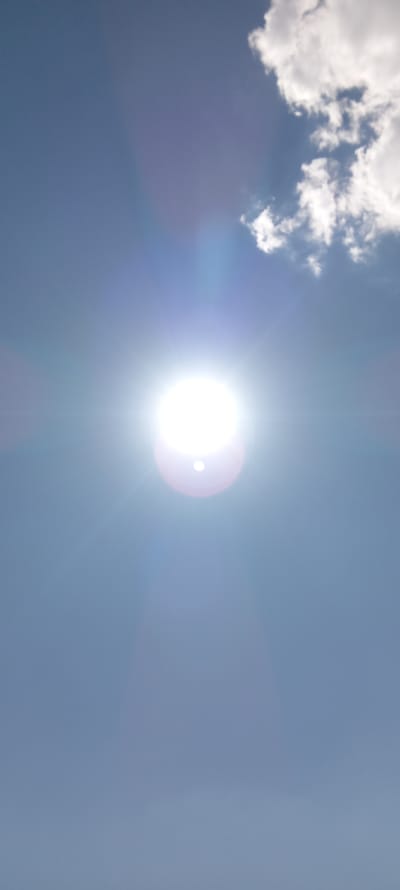 The image is a photograph of a bright sun in a clear blue sky with some white clouds visible in the upper right corner. The sun is positioned in the center of the frame and appears to be the main subject of the photograph, with prominent light flares emanating from it. The sky occupies the majority of the frame, with the clouds taking up a small portion of the upper right quadrant. There are no people, subjects, or objects other than the sky, sun, and clouds visible in the image. The photograph was taken outdoors, likely during the daytime given the presence of the sun. No specific location in the city of Juvarhas, Uzbekistan, can be identified from this image as it only depicts the sky.