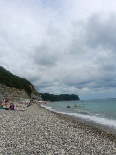 The image depicts a crowded pebble beach on a cloudy day, situated in Novomikhaylovsky, Russia. The beach is lined with lush green trees and layered rock formations on the left, with the turquoise sea on the right. Several people are scattered across the beach, sunbathing, walking, or sitting. In the water, a group of people are on a paddleboard, with two individuals visible. Some parked cars are visible along the road behind the beach. Umbrellas and beach chairs are set up in some areas, suggesting a recreational setting. The sky is overcast with a mix of dark and light gray clouds.