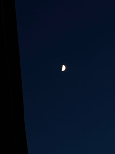 The image displays a dark blue night sky with a visible crescent moon. On the left side of the image, a dark, vertical object, possibly a tree or a building structure, occupies a significant portion of the frame. The moon is illuminated and appears in the upper right quadrant of the sky. The scene is devoid of any human presence or activity. The location is identified as Krasivka, Ukraine.
