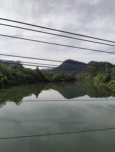 The image shows a landscape of a body of water surrounded by lush green hills and vegetation, likely a river or lake, in Lamseunia, Indonesia. The water's surface reflects the sky and the surrounding hills, creating a mirrored effect. Several black wires, possibly electrical or telephone, are strung across the top portion of the image. A tall, thin tower with red and white markings is visible on one of the hills in the background. The sky is overcast with a mottled pattern of clouds. No people are visible in the frame.