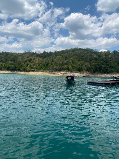 A landscape photograph shows a body of water next to land with dense foliage. The photo is taken on its side, with the land on the left and water on the right. The water is rippled and light blue. On the left is a sandy shoreline and thick green vegetation. A small boat is anchored near the shore, next to a small dock. Overhead is a blue sky with white clouds. The location is Ban Ta Khun, Thailand.
