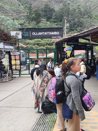 People wait at the Ollantaytambo train station. A sign above indicates the station name.  Nearby, a food stand advertises pizzas. A smaller booth offers coffee and ice cream. Patrons are seen boarding or queuing, some with backpacks. A money exchange booth is also present. The backdrop features a lush, green mountain.