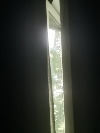 The image depicts a view through a narrow opening, possibly a gap between curtains or blinds, revealing a bright window. Sunlight streams through the window, illuminating what appears to be foliage outside, suggesting it's daytime. The immediate surroundings of the opening are dark, creating a strong contrast with the bright interior of the window. The scene appears to be indoors, with the dark areas suggesting walls or furniture covered by curtains. The location is not specified in the image itself or provided in the prompt.