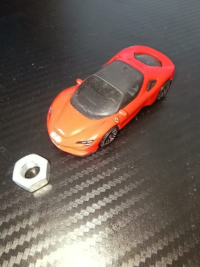 This image shows a red toy sports car and a metal nut lying on a textured, dark surface. The surface appears to be a carbon fiber pattern, commonly used in automotive aesthetics. The toy car is oriented with its front pointing towards the bottom right of the frame, and its body is predominantly red with black accents for the roof and windows. A small yellow and black emblem, likely a brand logo such as Ferrari, is visible on the side of the car. The metal nut is hexagonal and silver-colored, positioned to the right and slightly below the toy car. The lighting in the image suggests an indoor setting, possibly a desk or tabletop, with light illuminating the scene from the right, casting subtle shadows. There is no text visible in the image. The overall composition is a still life arrangement of these two objects.