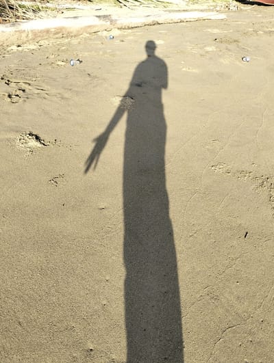 A shadow of a person is cast on a sandy surface. There are a few small cylindrical objects scattered across the sand. The background includes vegetation and a light-colored object, possibly driftwood. This photo was taken in Sagoe, Indonesia.