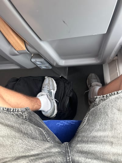 This image shows a close-up view of a person's legs and feet, with some surrounding objects. The person is wearing grey denim shorts and white socks with grey athletic shoes. Their right leg is bent, with the foot resting on a black duffel bag. A blue rectangular object, possibly a book or mat, is visible beneath the person's left leg and is partially obscured by their shorts.
The setting appears to be an indoor space, possibly a train or bus compartment, given the confined nature of the area and the presence of a white wall panel on the left and a dark floor. A gray electrical outlet with two ports is mounted on the white panel. A small piece of light-colored wood is positioned beneath the hinge of the wall panel. The lighting suggests it is daytime, but no specific time or weather conditions can be determined. The image is taken from a high-angle perspective looking down at the person's lower body. There is no visible text in the image.