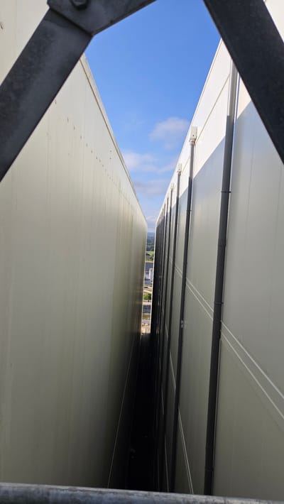 The image captures a view looking down a narrow gap between two tall, light-colored, paneled structures. The gap extends into the distance, where it meets a bright sky with scattered clouds. The structures have a uniform, ribbed texture on their exterior surfaces and appear to be industrial or commercial buildings. Along the right-hand structure, dark vertical pipes are visible, running from top to bottom and attached at intervals by brackets. The foreground shows a dark metal railing, suggesting the viewpoint is from an elevated position, possibly a walkway or platform. In the far distance, a town or rural landscape with greenery and some buildings can be seen through the opening. The scene is characterized by the stark contrast between the imposing verticality of the buildings and the open sky. The location of Écouché-les-Vallées, France, is not directly discernible from the visual elements of the image itself.