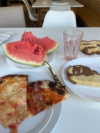 A close-up overhead shot shows a white table laden with food. In the foreground, two slices of pizza sit on a white plate. The pizza on the left is topped with melted cheese and tomato sauce. The pizza on the right has tomato sauce, cheese, and visible pieces of cured meat and olives. To the right of the pizza, another white plate holds three slices of bread spread with a chocolate hazelnut spread. A pink glass filled with water stands behind these plates. To the left of the pizzas, on a separate white plate, are several slices of watermelon, some of which have been bitten into. A knife with a wooden handle lies on the table between the watermelon and the chocolate bread. In the blurred background, white chairs and wooden tables suggest a dining area, possibly a restaurant or cafe. The scene is set in Dedovo, Bulgaria.