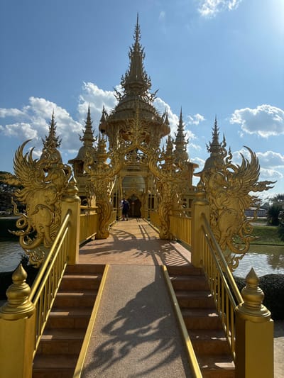 A golden, ornate structure stands tall against a blue sky with scattered white clouds. It features intricate carvings and decorative spires. The structure is connected to a walkway with golden railings and steps. Shadows stretch across the walkway's textured surface. The surrounding area includes water and greenery.