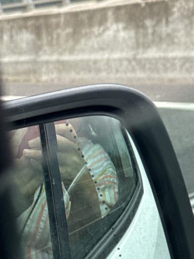 The image shows a reflection in a car's side mirror. Inside the reflection, a person is visible wearing a striped shirt with dark buttons. The person's hands are also visible, one holding a red object. The background in the mirror shows the interior of a car and the exterior environment, which appears to be a road or highway with a concrete barrier. The setting is the city of Carrigcloney, Ireland.