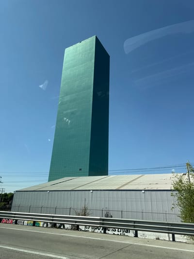 A tall, rectangular, green-colored building with a gridded facade is seen against a clear blue sky. Part of the sky has a reflection on the building. To the right of the building is a large, metal-sided industrial building or warehouse with horizontal ridges, also seen from a low angle. A highway or road runs along the right edge of the image, with a metal guardrail. There is graffiti visible on the guardrail and the adjacent wall of the industrial building. Vegetation can be seen near the base of the industrial building and at the very top of the image. The location cannot be confirmed as Santa Ana Xalmimilulco, Mexico based solely on the visual information provided.