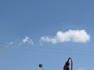 The image depicts a clear blue sky with a few scattered white clouds. Two thin, dark power lines stretch horizontally across the frame. In the lower right corner, a red, stylized object resembling a vintage lamp or speaker is partially visible. To the left of this object, a portion of another similar object, possibly a lamp with a circular top, is also visible. The scene appears to be outdoors, with the sky dominating the background. The overall impression is one of a bright, possibly sunny day. There are no people visible in the image, and no specific landmarks of Kyzyltepa, Uzbekistan, are identifiable.