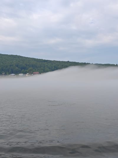 A wide shot captures a hazy, overcast day over a body of water in Atamanovo, Russia. The water's surface is rippled, reflecting the muted sky. In the distance, a dense green forest covers a rolling hillside. A low-lying mist obscures the base of the hill and creeps out over the water, partially concealing a small cluster of houses nestled along the shore. The houses appear to be simple structures, some with visible roofs. The overall atmosphere is serene and somewhat mysterious due to the fog.