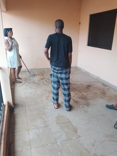 In Ufuma, Nigeria, an indoor or semi-enclosed space with light peach-colored walls and light-toned tiled flooring is depicted. The floor is partially covered in water and soap suds, indicating an active cleaning process.

Three individuals are present. On the left, a female subject with light skin wears a light-colored sleeveless dress and a dark head covering. She holds a wooden-handled mop with a circular head, resting on the wet floor. Her head is turned towards the right, and her left hand is raised slightly, fingers together. She wears light-colored flip-flops.

In the center, with his back mostly to the camera, stands a male subject with dark skin. He wears a black short-sleeved t-shirt, blue and white plaid pants, and dark flip-flops. His left hand is placed in his pants pocket. A dark green bucket rests on the floor near his feet. He is oriented towards the right side of the frame.

On the far right, only the lower leg and foot of a third subject are visible, wearing a dark blue Crocs-style slipper.

The background on the right features a wall with a dark-framed rectangular window, possibly fitted with louvers or a security screen. The foreground on the left includes a dark vertical bar, potentially part of a railing or window frame.
