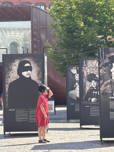 In Lund, Sweden, on a sunny day, an outdoor exhibition displays large black-and-white portraits of individuals in historical attire. One prominent portrait shows a person with their eyes covered by a black band. A woman wearing a red dress stands in front of the portraits, shielding her eyes from the sun as she observes the exhibition. The background reveals elements of Lund's urban landscape, including a modern building with a dark facade and a glimpse of a historical brick building with arched windows, likely St. Peter's Abbey, a significant landmark in Lund. The ground is paved with light-colored cobblestones, casting distinct shadows from the structures and the exhibition panels.