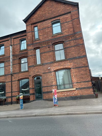 A three-story brick building is depicted in this image, located in Crewe, United Kingdom. The building has a dark, sloped roof and numerous windows with dark frames. A vertical banner with white text reading "HERE FOR YOUNG PEOPLE" is attached to the left side of the building. Two signs are placed on the sidewalk in front of the building; one is blue and advertises food with the text "Savour Every Bite" and "Try our Nation's Chicken." The other sign appears to be a colorful advertisement for food items like ice cream. The building is situated next to a paved walkway and a street. The sky is overcast with clouds. There are no people visible in the image.