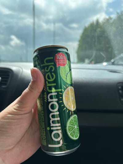 In Arkhangelskoye, Russia, a hand holds a green aluminum can of "Laimon Fresh" beverage. The can is adorned with images of lime and lemon slices, along with mint leaves. A red circular label on the can reads "100% НАТУРАЛЬНЫЙ СОСТАВ" (100% Natural Ingredients). The background suggests the can is inside a vehicle, with the cloudy sky and trees visible through the windshield. The interior of the car, including the dashboard and controls, is also partially visible.