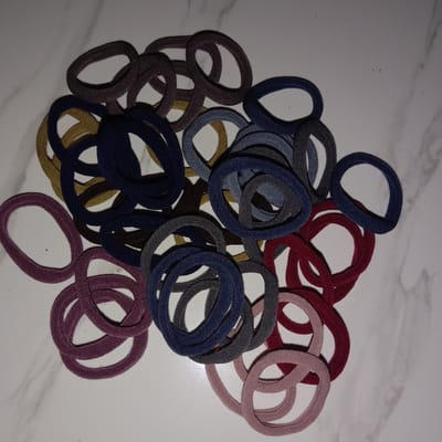 A pile of hair ties with various colors (purple, brown, gold, dark blue, grey, red, light pink) on a white marble-like surface. The location is unspecified and the image does not suggest it is in Jatisari, Indonesia. There are no people in the image.