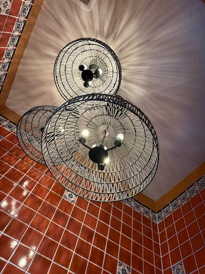 The image depicts an upward view within the interior of Miguel's Cocina in Chula Vista, United States of America. Three large, circular ceiling light fixtures dominate the frame. Each fixture consists of a dark metal framework with multiple exposed incandescent bulbs, surrounded by a wide, woven shade crafted from light-colored cord or yarn wrapped around a dark circular rim. The uppermost fixture casts distinct radial light patterns onto the pale, textured ceiling. The upper portions of two interior walls are visible, meeting at a corner. These walls are clad in uniform reddish-brown square tiles with light-colored grout lines. A decorative border, featuring small patterned blue and white tiles topped by a solid ochre-yellow band, runs along the junction of the tiled walls and the light-colored ceiling. No individuals, actions, or interactions are present in the scene.