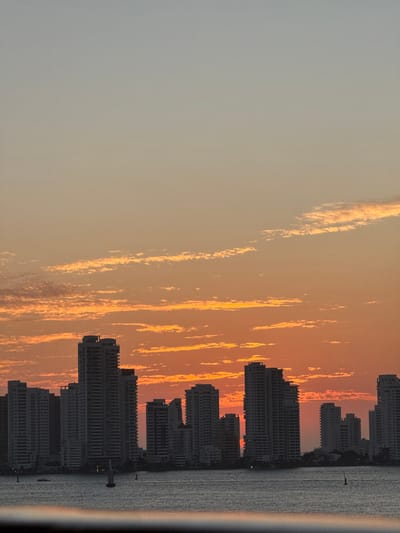 A cityscape silhouette against a vibrant, striated sky is depicted. The sky transitions from a soft grey to a warm orange with streaks of yellow. Tall buildings create a jagged, dark outline along the horizon. Water is to the right of the buildings.