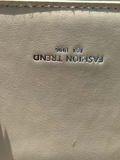 The image shows a close-up of a beige textured surface, possibly leather, with embossed silver lettering. The lettering reads "FASHION TREND EST 1996". The surface is lit from the side, creating shadows that highlight the texture. The top edge of the image shows a seam with visible stitching, suggesting this is part of a bag or accessory. There are no people or any specific environmental details visible to indicate a location within Парыцкі сельскі Савет, Belarus.