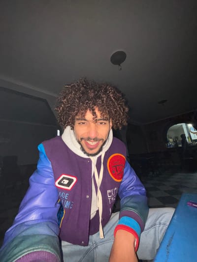 A man with curly hair and a beard smiles broadly, wearing a white hooded sweatshirt and a purple jacket with blue sleeves. The jacket has patches, including a face and a geometric design. The background shows a dark room, a checkered floor, and dim lighting. A blue table is visible on the right. The perspective is from a low angle.