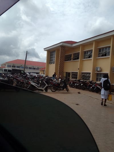 The scene depicts an outdoor setting in what appears to be Orifite, Nigeria, given the architectural styles and general environment. The foreground includes a dark-toned obstruction, likely part of a vehicle interior, partially obscuring the view. Midground shows numerous parked motorcycles and some vans in a parking lot. Buildings with yellow facades and red roofs are present. A person in a white uniform and carrying a yellow bag walks on a brick-paved walkway. A person is standing near the parked motorcycles. The sky is overcast. Electrical wires are visible in the background.