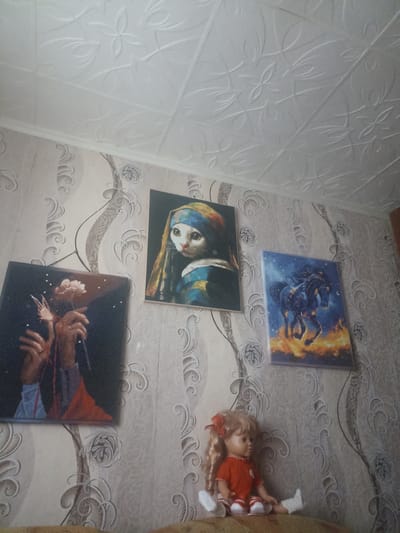 The image depicts an indoor scene, likely a room in a residential building. The walls are covered in patterned wallpaper, and the ceiling is textured with a repeating floral motif. Three framed pictures are hung on the wall. The leftmost picture shows hands holding a light-colored flower against a dark background, with small white dots appearing like stars. The middle picture is a diamond painting of a cat with a blue and yellow headscarf, resembling "Girl with a Pearl Earring." The rightmost picture is a diamond painting of a black horse running through flames against a starry blue sky. In front of the wall, a doll with blonde hair, wearing a red dress and white socks, is seated. The overall impression is that of a decorated living space. The location is identified as Perelešinsky, Russia, suggesting this is a snapshot of a home in that specific area.