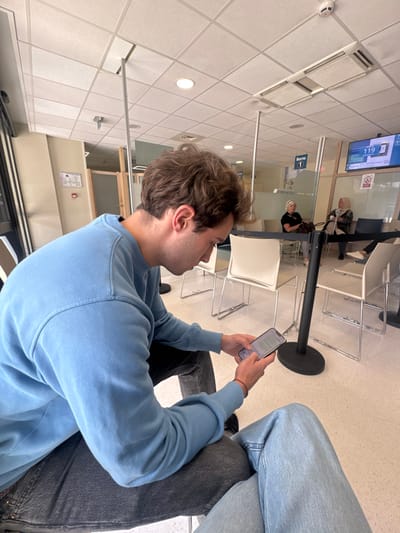 The image depicts an indoor waiting area within a public service office in Saint-Priest, France. In the foreground, a male individual, approximately 20-30 years old with light brown, wavy hair, is seated. He wears a light blue crew-neck sweatshirt and dark grey jeans, and is focused on a light-colored smartphone held with both hands, interacting with the screen. A thin red string bracelet is on his right wrist. The leg of another person, wearing light blue jeans, is partially visible in the bottom right.

The waiting area features a grid-patterned suspended ceiling equipped with recessed circular lighting fixtures, rectangular air vents, and a smoke detector. The floor is a light-colored, speckled tile or linoleum. Transparent glass partitions with vertical metal supports delineate sections of the space. Black stanchions with retractable belts are present for queue control.

In the mid-ground, two women are seated in white, contemporary-style chairs with metal frames. One older female, with blonde hair and a black top, is positioned center-left, with a dark bag on an adjacent chair. Another female, wearing a headscarf and a patterned top, is seated further right.

On the far right wall, a digital queuing system display shows "119" as the current number being served, with an associated counter "25" indicated below. A red circular 'No Smoking' emblem is affixed to the wall beneath the display. A sign marked "Borne 1" (Kiosk 1) is mounted above a glass partition. A red-bordered informational sign is affixed to the left wall. The scene suggests a typical public administrative or service waiting room environment.