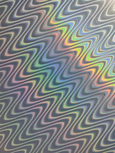 The image shows a close-up of a holographic surface with a repeating wavy pattern. The base color appears to be a light gray, but the holographic effect produces iridescent bands of color that shift between greens, blues, pinks, and yellows. The light source is not directly visible, but the reflection and refraction of light off the surface create a dynamic and colorful visual effect. The surface appears to be slightly reflective and has a smooth texture. No people, specific subjects, objects, or locations in González Catán, Argentina are discernible in the image.