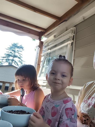 An outdoor, waist-up shot captures two young girls and a partial view of a third person, possibly an adult, during a meal. The scene appears to be on a covered porch or patio. The girl on the left, with shoulder-length brown hair, wears a pink tank top and is looking down, holding a spoon in a blue bowl filled with dark berries. She is positioned in front of a white bowl. The girl on the right, with her hair in two buns, is facing the camera and smiling. She is wearing a light pink shirt with a strawberry print and is also in front of a blue bowl of berries. Part of a person in a yellow shirt with a cartoon print is visible on the far right. The background includes a beige, horizontal-paneled wall, a white-framed window, and wooden beams supporting the roof structure. A tall, dark green tree is visible against a bright sky in the distance, along with a white mobile home or trailer. The overall lighting suggests it is daytime, likely sunny.
