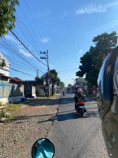 The image depicts a street scene in Margoyoso, Indonesia, under a clear blue sky. A person is riding a red motorcycle in the foreground, facing away from the camera and wearing a pink helmet and a green and black outfit. The motorcycle has a license plate reading "K 4934 UBA". The street is paved with asphalt and lined with gravel on the sides. To the left of the street are buildings with blue metal fences and some greenery. To the right, more buildings and trees are visible, with a dense canopy of leaves from a large tree dominating the right side of the frame. Power lines are strung across the sky, supported by utility poles. In the distance, other motorcycles can be seen on the road. The sun is casting shadows, indicating it is likely daytime. The image appears to be taken from the perspective of someone on another motorcycle, with the edge of a helmet visible in the bottom right corner.