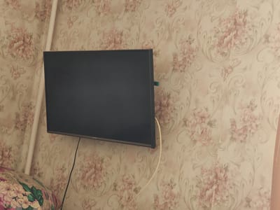 A wall-mounted flat-screen television is the main subject, centrally positioned against a backdrop of floral wallpaper. The wallpaper features a repeating pattern of pink and cream-colored flowers with scrolled green and beige accents. To the left of the television, a vertical white pipe, likely a radiator pipe, runs from top to bottom. A cream-colored cable extends from the right side of the television, looping downwards and then disappearing out of frame. On the lower left side of the image, a portion of a patterned cushion or piece of fabric is visible, with a floral design featuring pink and green elements. The television itself is dark and reflects some light from the environment. On the bottom right edge of the television, the word "SAMSUNG" is visible in small white text. The lighting suggests an indoor setting, possibly during the daytime. There are no people visible in the frame, and no specific activity or event is taking place. The overall scene depicts a typical living space, with a focus on the electronic device and its placement on the wall.