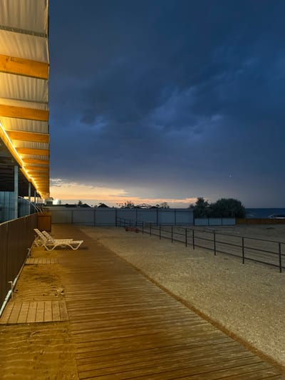 The image depicts a beachfront area at dusk or early evening. On the left, a wooden boardwalk runs parallel to a brown wooden fence. Two white lounge chairs are positioned on the boardwalk. Above the boardwalk, a covered structure with yellow lighting runs along its length. The ground beyond the boardwalk is covered in sand and small pebbles, bordered by a black metal fence. In the distance, there are scattered buildings, trees, and the sea. The sky is dark blue with some clouds, and a faint hint of orange and pink from the setting sun is visible near the horizon. A small, red wheeled cart is visible on the sandy area. A single white speck, possibly a distant light or star, is present in the sky. The scene appears to be located in Izberbash, Russia, based on the provided location context.