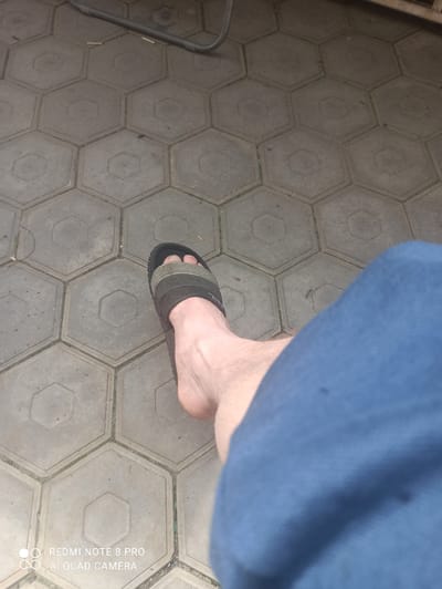 The image shows a person's foot wearing a black and green sandal, resting on a paved surface composed of hexagonal concrete tiles. A portion of blue fabric, likely clothing, is visible on the right side of the frame, obscuring part of the person's leg. In the background, a dark, curved object, possibly part of a piece of furniture or equipment, is visible. The scene appears to be outdoors, on a ground-level surface. The image is watermarked with "REDMI NOTE 8 PRO AI QUAD CAMERA" at the bottom left. The specific location in the city of Irbit, Russia cannot be determined from the visual information provided.